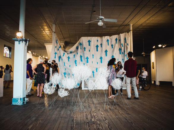 Several people stand in a large room. Artwork is hung throughout. Sculptures that recall dandelion seeds are arranged in the foreground. A large tarp painted with upward-pointed arrows hangs in the background.