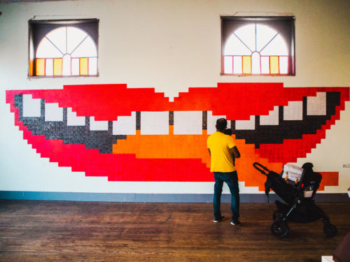 A man holding a baby looks at a large mural painted on an interior wall. The mural looks like a mouth with red lips. In this context, the windows above the mural look like eyes.