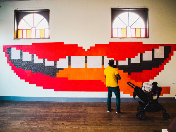 A man holding a baby looks at a large mural painted on an interior wall. The mural looks like a mouth with red lips. In this context, the windows above the mural look like eyes.