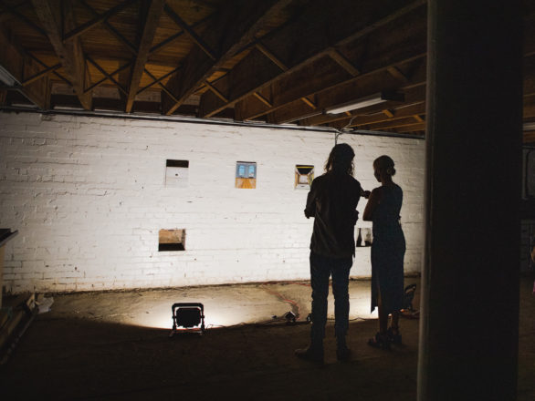 Two people look at paintings hung on a brick wall. The ceiling is low, and the floor is rough. Spotlights illuminate the wall in the otherwise dark room.