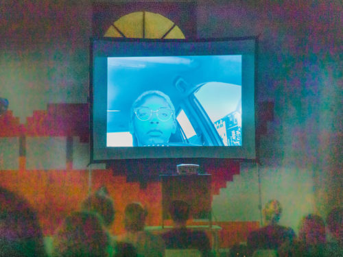 An image of the face of a young Black woman is projected onto a screen. Audience members are looking up at the screen.