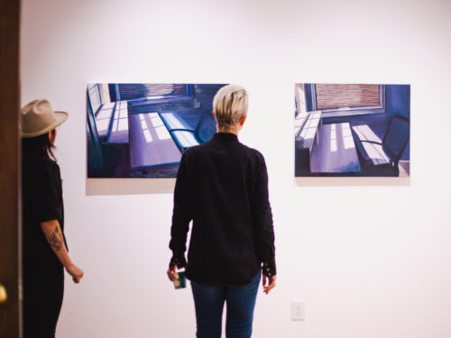 Two women stand in an art gallery, facing a wall upon which two medium-sized paintings hang.