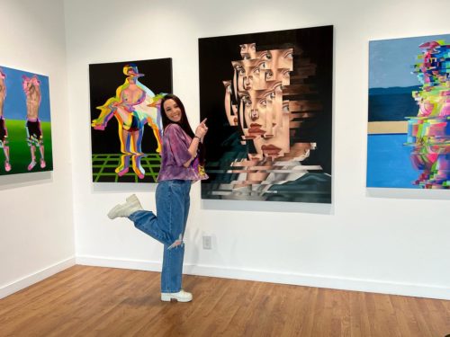 A woman poses in an art gallery, smiling and pointing at one of her paintings.