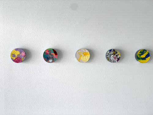 Five small paintings hanging on a wall. The objects are circular, and are made by applying paint and collage to the bottoms of tuna cans.