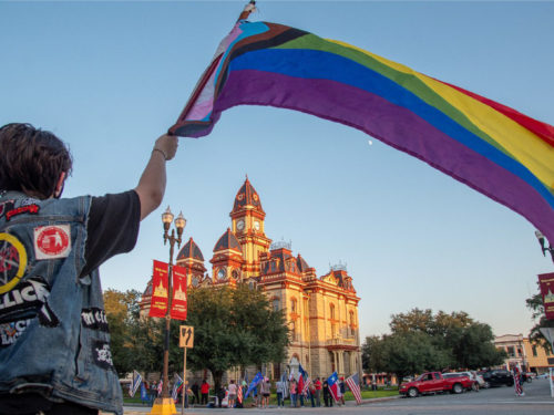 A person wearing a jean jacket waives a pride flag, where across the street a crowd has gathered with American and Texan flags on the grounds of the Caldwell County courthouse.