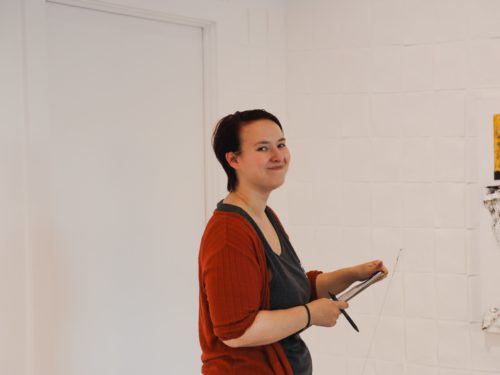 Alex Renbarger, a woman standing in a gallery with a clipboard. She is wearing a red cardigan.