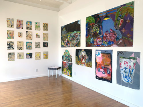 Artwork on view in an art gallery. On one wall, twenty small drawings hang on a grid. Seven larger paintings hang on another wall. The style is chaotic and dark.